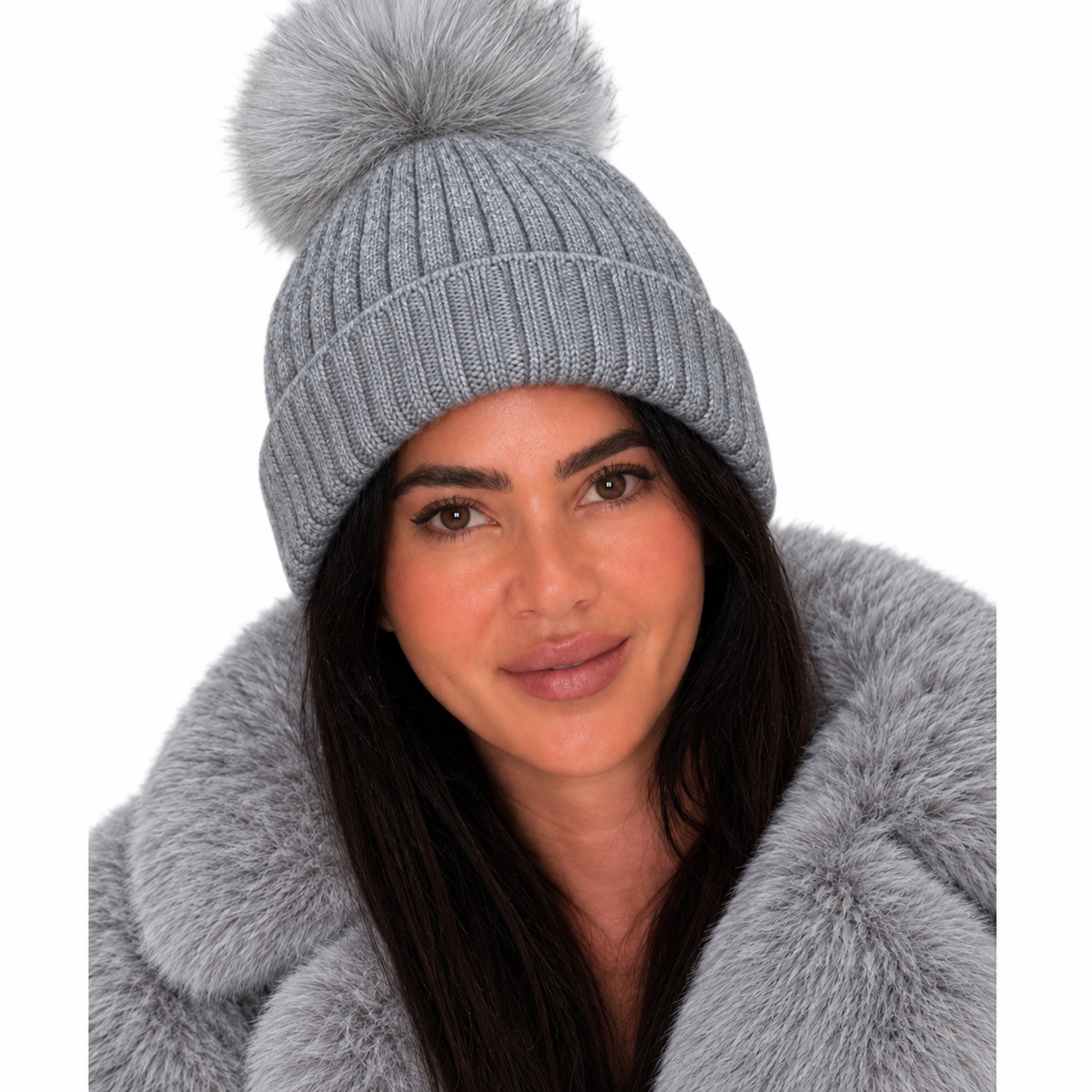A woman wearing a light gray ribbed knit beanie with a large fluffy pom-pom, paired with a matching faux-fur collar, against a plain white background.
