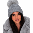 A woman wearing a light gray ribbed knit beanie with a large fluffy pom-pom, paired with a matching faux-fur collar, against a plain white background.