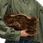 A person in an olive green shirt holds a brown faux fur clutch bag with a tan handle and zipper.